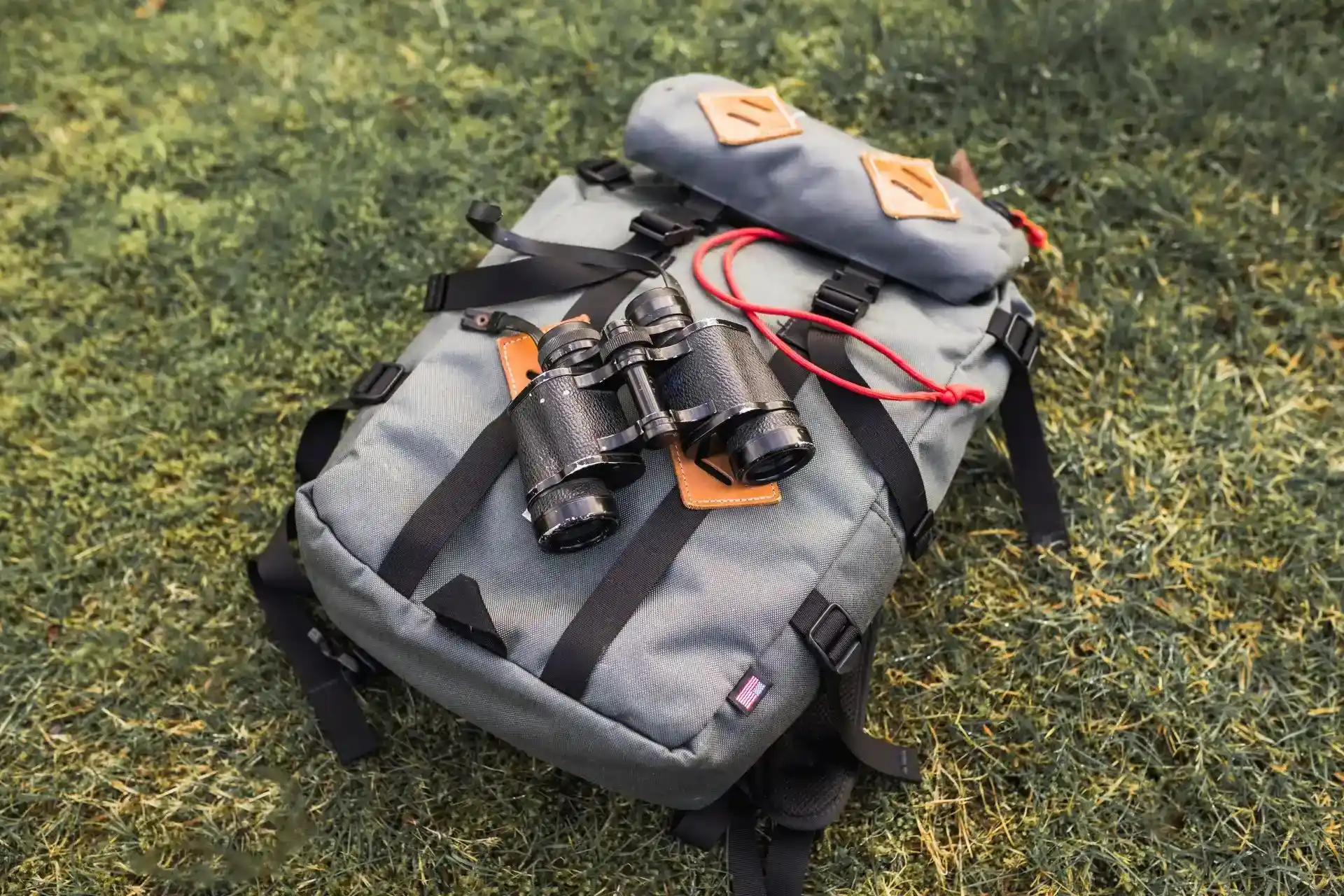 Sac à dos gris et noir posé sur l'herbe, équipé de jumelles et de diverses sangles et accessoires